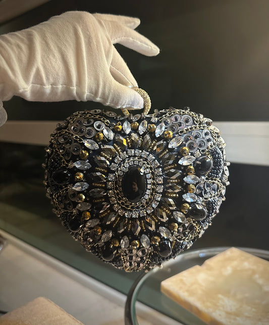 Clutch en forma de corazón con embargue de diferentes tipos de piedras base color negro, dorado y plata.